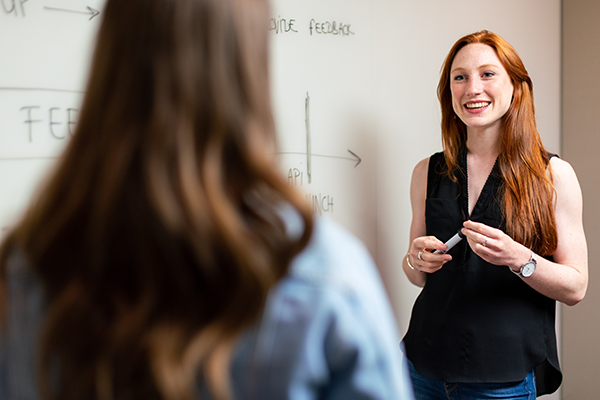 Young teacher standing next to a board, smiling and speaking to a student.
